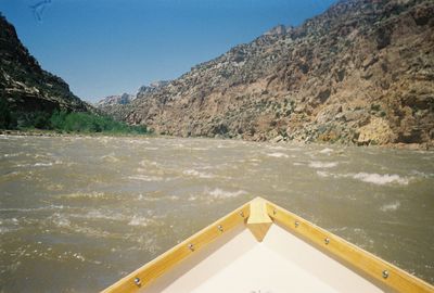 The bow of a wooden dory points downstream into wind-chopped brown water in Split Mountain Canyon, with tan tilted canyon walls under a clear sky.