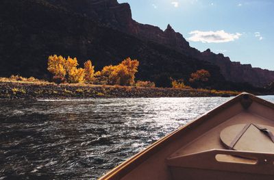 Backlit golden cottonwoods glow along a cobble bank of the Green River, seen over the bow of a dory, with a shadowed canyon wall of Split Mountain rising behind.