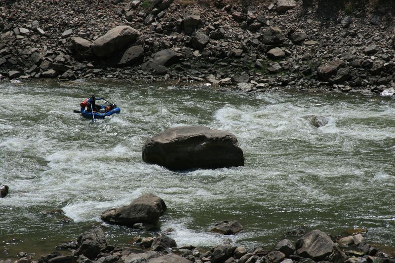 A rower in a loaded blue raft threads a rocky rapid on the Green River, pulling past a large mid-channel boulder amid whitecaps.