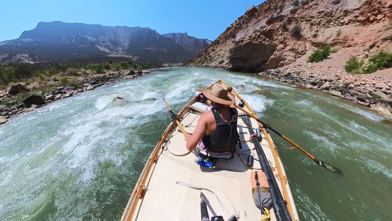 Bow-forward fisheye view from a wooden dory running a low-water wave train in Split Mountain Canyon on the Green River, the straw-hatted rower pulling on the oars between dry tan-and-red canyon walls.
