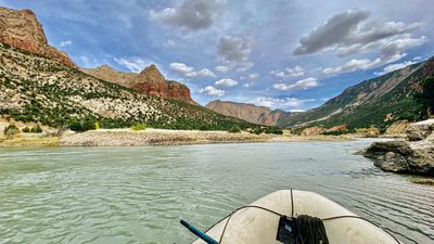 The bow of a tan raft with a coiled bow line and an oar shaft crosses calm green water toward the red crags and distant gates of Split Mountain under a dramatic cloud-filled sky.
