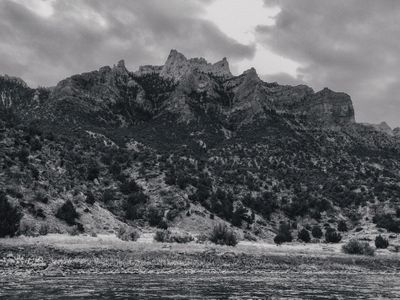 Black-and-white view of the jagged crest of Split Mountain under heavy clouds, above juniper slopes, a pale grass bench, and the textured surface of the river.