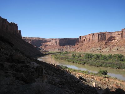 Morning view down Stillwater Canyon as the Green River winds between green riparian benches and red sandstone walls, with a shadowed talus slope in the left foreground.