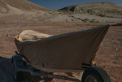 A dory under a tan canvas cover sits on its trailer on bare desert ground, pale eroded badland hills rising behind it in low warm light.