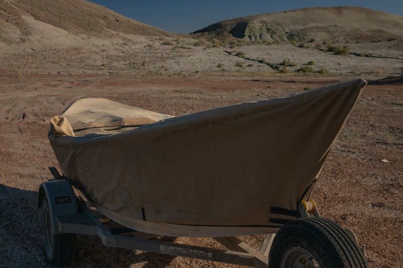A dory under a tan canvas cover sits on its trailer on bare desert ground, pale eroded badland hills rising behind it in low warm light.