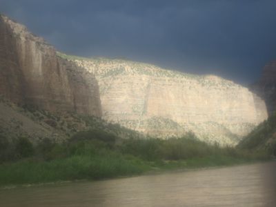 Storm light breaks across the tall layered wall of Whirlpool Canyon above green riverside vegetation on the Green River, with rain haze softening the frame under a dark sky.