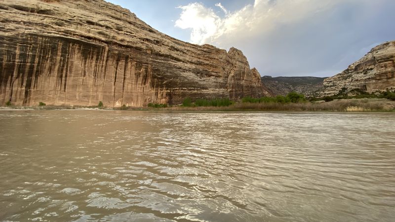 Massive desert-varnish-streaked sandstone wall rising straight from the silty river as sunlight breaks through clouds above, seen from water level.