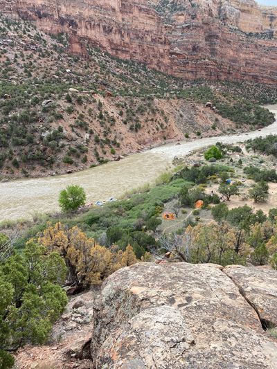 Looking down from a rocky viewpoint at a river camp on a juniper bench, tents scattered in sandy clearings and boats tied along the willow-lined bank below red-brown canyon walls.