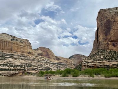 A single oar raft carrying three people across wide flat water, dwarfed by a sheer sandstone cliff on the right and rounded slickrock domes upstream.