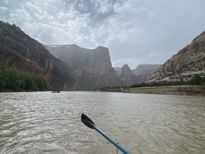 View from a boat in falling rain, a blue paddle in the foreground and a yellow raft far ahead on gray water beneath dark, rain-streaked canyon walls.