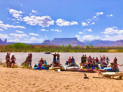 A large multi-family river group posed together on a sandy beach beside their loaded rafts and inflatable kayaks on the Colorado River, red-rock mesas and the snow-capped La Sal Mountains rising under a bright blue sky behind them.
