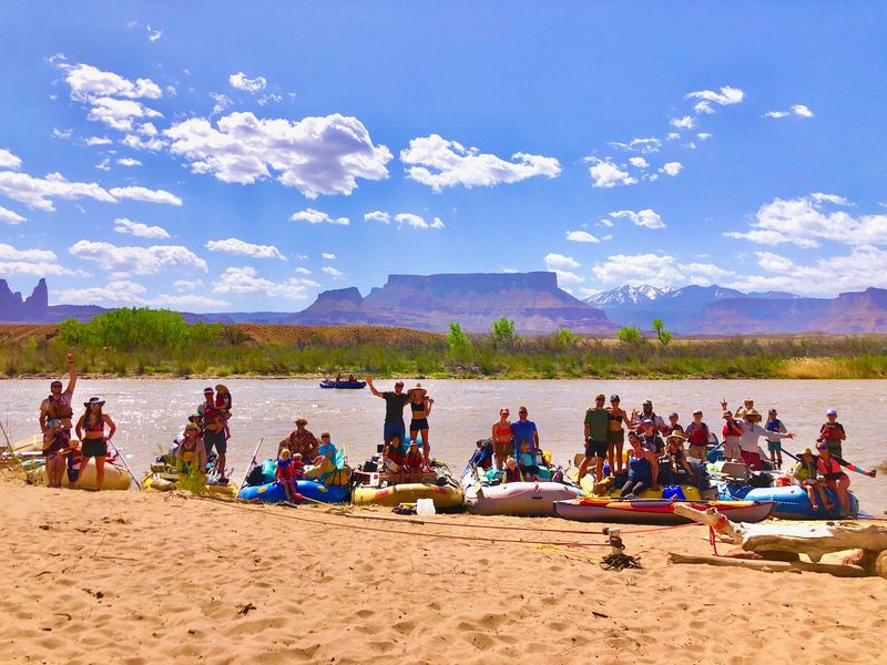 A large multi-family river group posed together on a sandy beach beside their loaded rafts and inflatable kayaks on the Colorado River, red-rock mesas and the snow-capped La Sal Mountains rising under a bright blue sky behind them.