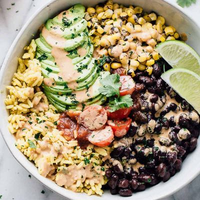 Overhead view of a white bowl filled with yellow rice, black beans, charred corn, sliced avocado, cherry tomatoes, and salsa drizzled with a creamy sauce, garnished with cilantro and lime wedges.