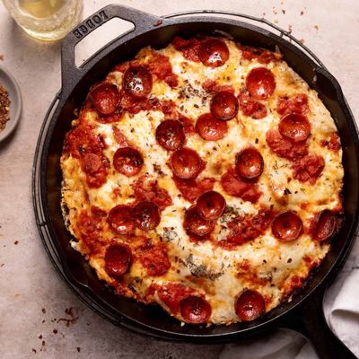 Overhead view of a pepperoni pizza baked in a black cast-iron skillet, its cheese browned and bubbled, with chili flakes and a small dish of seasoning on the table nearby.