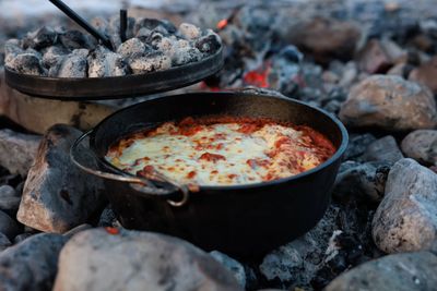 A cast-iron dutch oven full of bubbling, cheese-topped lasagna sits among gray river cobbles and glowing embers, its coal-covered lid lifted to one side.