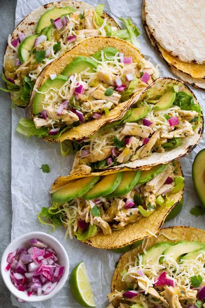 Charred corn tortillas filled with shredded salsa verde chicken, avocado slices, lettuce, shredded cheese, and diced red onion on parchment, with a small bowl of onion and a lime wedge alongside.
