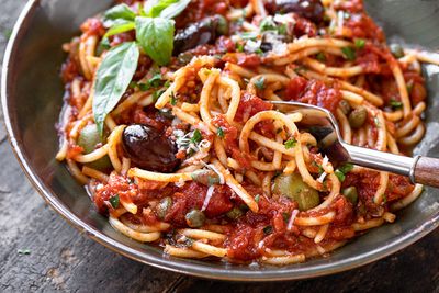 Close-up of a metal bowl of spaghetti puttanesca in chunky tomato sauce with olives and capers, garnished with fresh basil, on a dark wood table.