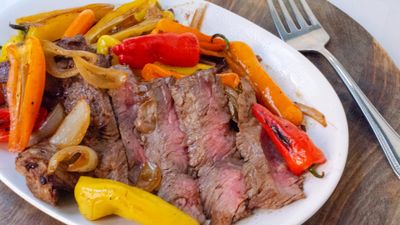 Slices of medium-rare ribeye on a white oval platter with blistered mini bell peppers and caramelized onions, a fork resting on the wooden board beside it.