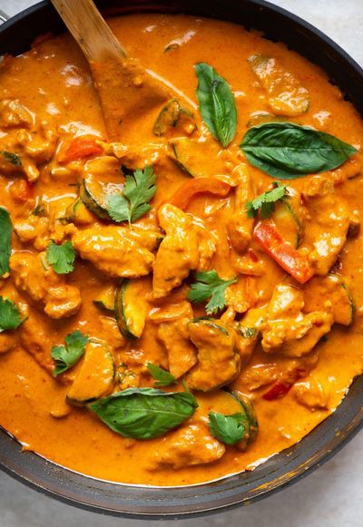 Close-up of a dark skillet of Thai red coconut curry with chicken, zucchini, and red bell pepper in orange sauce, garnished with basil and cilantro, a wooden spoon resting in the pan.