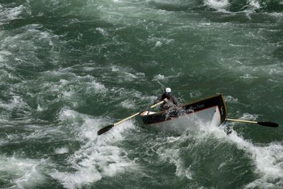 A rower pulls a wooden dory through heavy whitewater, oars braced and spray over the bow.