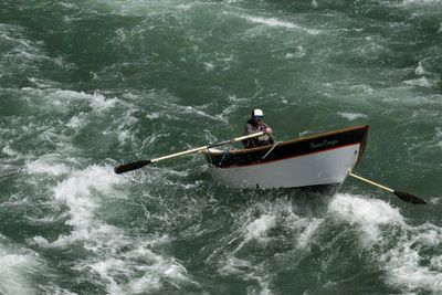 A rower holds a wooden dory steady in green whitewater, both oars out and spray along the hull.
