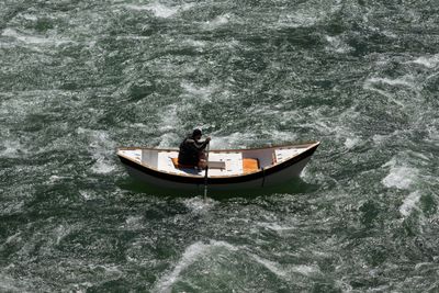 A wooden dory sits low in churning green water, a single rower at the oars.