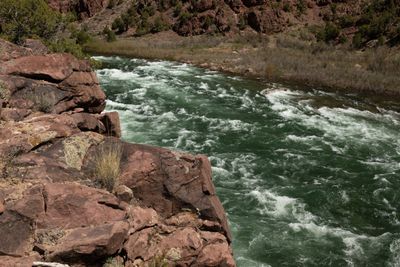 Looking down from a red rock ledge onto a green rapid running below juniper-dotted cliffs.