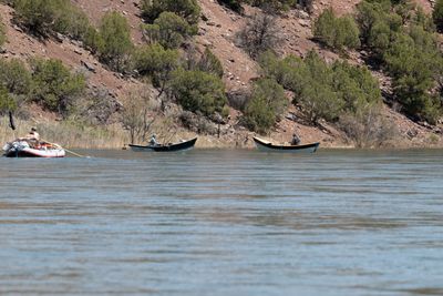 Three boats — a loaded raft and two dories — drift flat water below a juniper-dotted slope.
