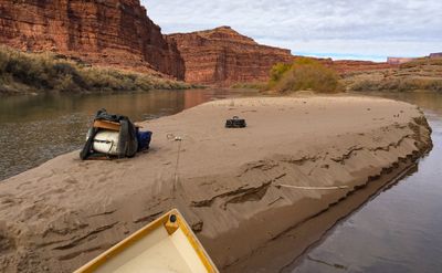 A dory's bow rests against a broad sand spit where a rolled bedroll, a wooden frame and a small hard case sit in the sand, with red sandstone cliffs and a brushy bank across the flat river.