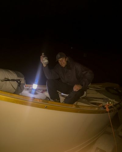 A crew member in a rain jacket and ball cap crouches in the open bow of a wooden dory at night, holding up a small bottle beside a lit battery lantern, with everything past the boat in darkness.