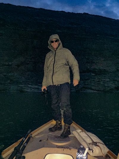 A crew member in a hooded jacket and sunglasses stands on the bow deck of a dory in near-darkness, a black canyon wall and slate-blue dusk sky behind, with oars and a rolled bag in the foreground.