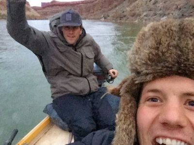 A close selfie of a grinning crew member in a fur trapper hat, with a second crew member behind him at the oars of a dory in a heavy hooded jacket, one arm raised, red canyon walls and cold gray-green water behind.