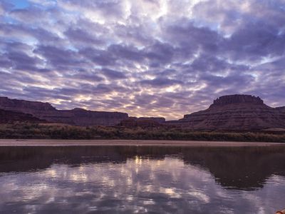 Broken altocumulus cloud with sun burning through low reflects across a wide, glassy stretch of the Colorado River at dusk, with dark silhouetted red-rock mesas and a pale sandbar along the far bank.
