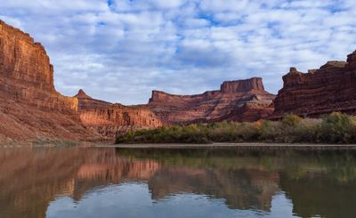 Sunlit red sandstone cliffs and a flat-topped butte rise above a yellowing willow bank, all reflected in the still gray-green Colorado River under a patchy cumulus sky.