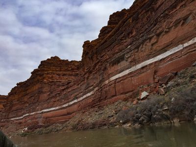 A steeply tilted red sandstone wall rising straight from the river with a bright white band of rock running through the layers, blocky rubble and dry brush along its base, under flat gray cloud.