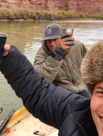 Two crew members in the dory raise metal cups toward the camera — one in a fur-ruffed hood in the foreground, the other in an olive jacket and ballcap — with brown water, a brushy bank and red cliffs behind under overcast sky.