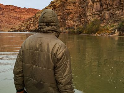 Seen from behind, a hooded crew member in an olive puffy jacket sits forward in the dory with gloved hands on the wooden gunwale, facing a glassy brown river running beneath a tall red sandstone wall under flat gray sky.