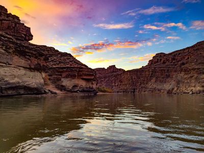 Dusk over the river: dark layered canyon walls stand in silhouette on both sides while a pale orange and blue sky reflects in long ripples across the water.
