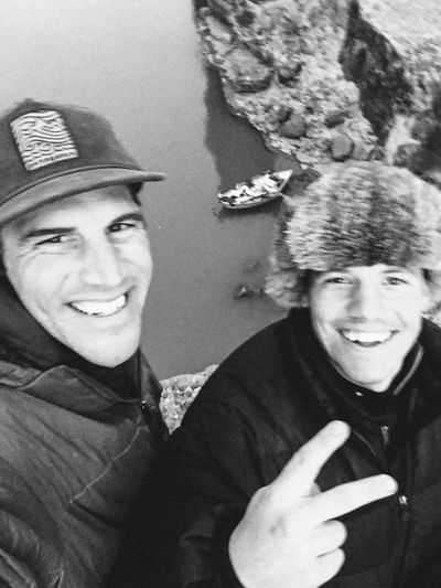 Two crew members grin in a selfie from a rock ledge above the river — one in a ballcap, one in a fur trapper hat flashing a peace sign — with the loaded dory tied off on the green-brown water below.