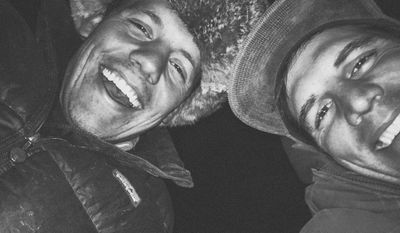 A night flash selfie of two crew members lying back and laughing, one in a fur trapper hat and one in a gray beanie, both in down jackets against full darkness.