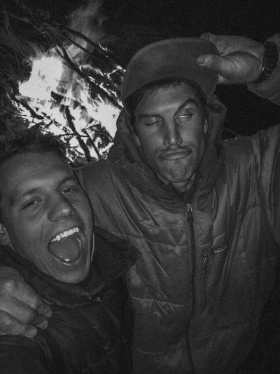 Two crew members mug for the camera in front of a large driftwood campfire at night, whole burning branches standing up in the flames behind them, both in hooded puffy jackets.