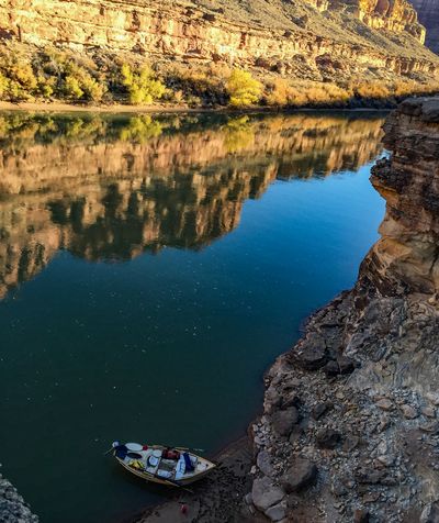 From a rocky ledge, a wooden dory is beached on a small sand and cobble landing while the reflection of a sunlit sandstone wall and yellow trees fills most of the still water.