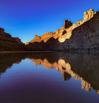 From inside a dory, calm dark water mirrors sunlit sandstone towers and buttes along the right-hand rim under a cloudless deep blue sky.