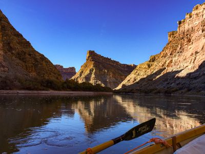 Looking downstream past an oar shipped on a dory's gunwale toward a sunlit layered butte standing above a river bend, with a low sandbar along the left bank.