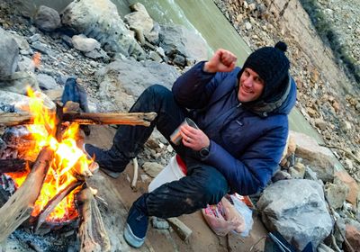 A crew member in a puffy jacket and knit beanie sits on the cobble beside a driftwood fire burning in a fire pan, holding an insulated mug, with the cold river running just behind.