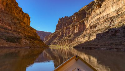 From the bow of a wooden dory, a calm river runs between tall sunlit red sandstone cliffs and shadowed talus slopes under a deep blue sky.
