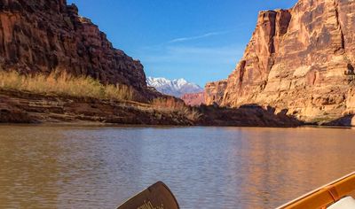 A distant range of snow-covered peaks shows through a downstream gap in the canyon, framed by red cliffs above a wide silt-brown river, with an oar blade in the foreground.