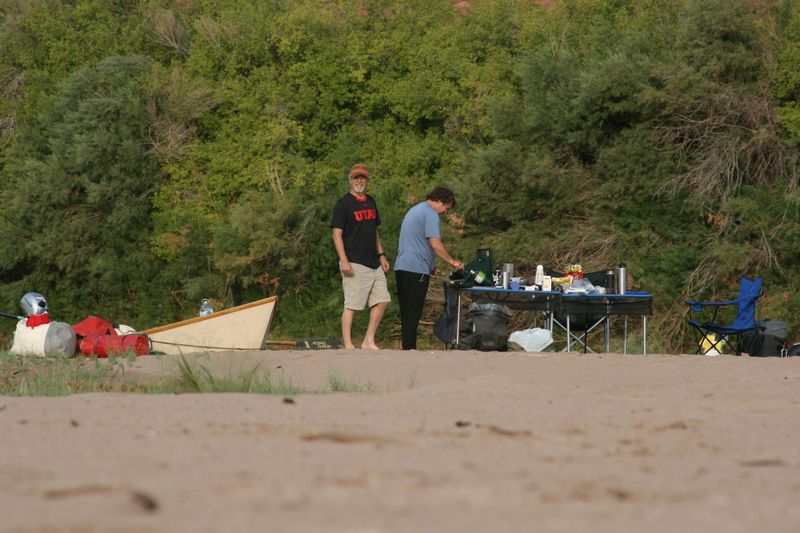 Two people stand at a folding camp kitchen table loaded with stove, cookware and food on a broad sand beach