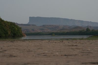 A flat stretch of river in dim dusk light with a hazy flat-topped mesa on the far skyline, low green brush along both banks and a broad rippled sand flat filling the foreground.