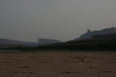 Dusk view over a dark sand flat toward a low silhouetted skyline of mesas and a single knob-topped butte, with two birds in flight against the pale gray sky.
